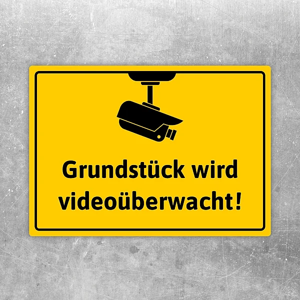 Gelber Aufkleber mit Kamera-Symbol und Text „Grundstück wird videoüberwacht“ für den sichtbaren Warnhinweis.
