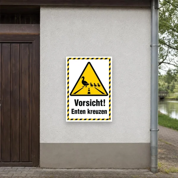 KI-generiertes Anwendungsbeispiel: Warnaufkleber Vorsicht! Enten kreuzen als Hochformat-Aufkleber an einem Weg nahe einem Teich.