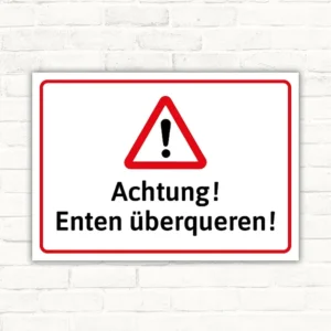 Warnschild mit dem Text Achtung! Enten überqueren! in Querformat mit rotem Warndreieck und schwarzer Schrift.