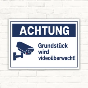 Schild mit Kamera-Symbol und Text „Grundstück wird videoüberwacht!“ als Hinweis für Haus und Einfahrt.