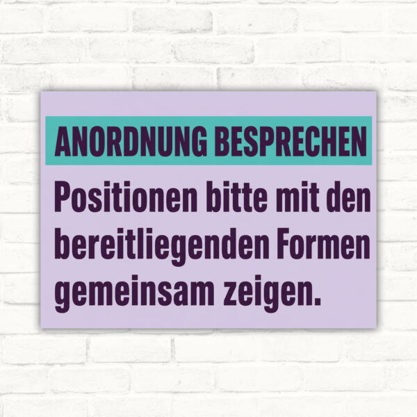 Schild „ANORDNUNG BESPRECHEN“ mit Text zu bereitliegenden Formen und zwei Personen beim Zeigen von Positionen.