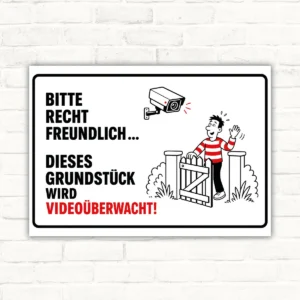 Schild mit Text „Bitte recht freundlich... dieses Grundstück wird videoüberwacht!“ und Kamera-Motiv als Hinweisschild.