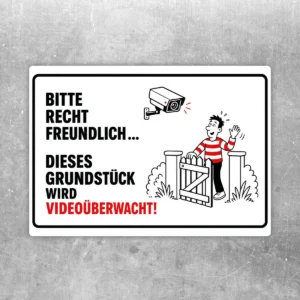 Freundlicher Aufkleber mit Kamera-Motiv und Text „Dieses Grundstück wird videoüberwacht“ in Schwarz, Rot und Weiß.