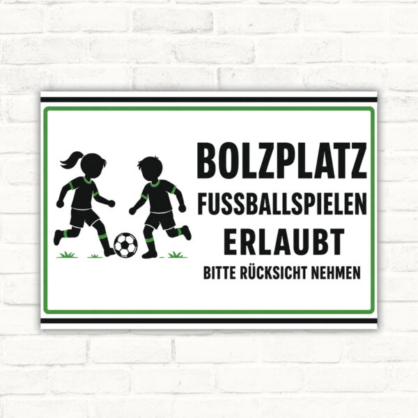 Bolzplatz-Schild mit Fußballkindern und Text „Fussballspielen erlaubt – bitte Rücksicht nehmen“ als Querformat.